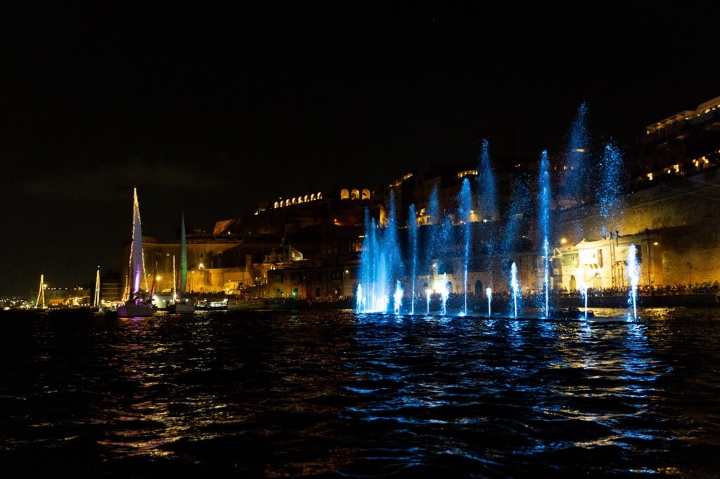 A photo showing Illuminated water fountains and sailing vessels during the Valletta Pageant of the Seas in Malta's Grand Harbour.