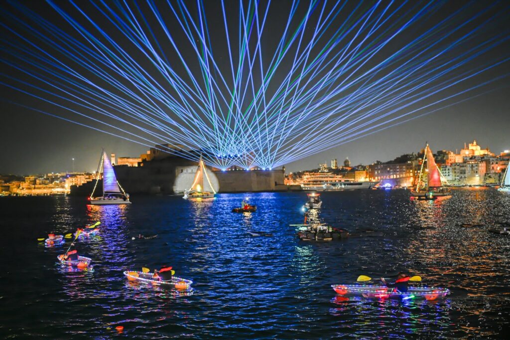 A photo showing Illuminated water fountains and sailing vessels during the Valletta Pageant of the Seas in Malta's Grand Harbour.