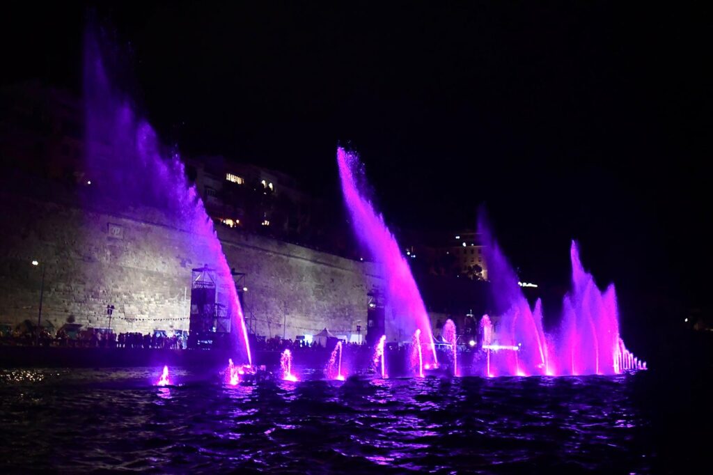A photo showing Illuminated water fountains and sailing vessels during the Valletta Pageant of the Seas in Malta's Grand Harbour.