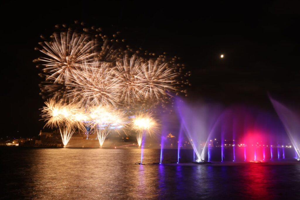 A photo showing Illuminated water fountains and sailing vessels during the Valletta Pageant of the Seas in Malta's Grand Harbour.