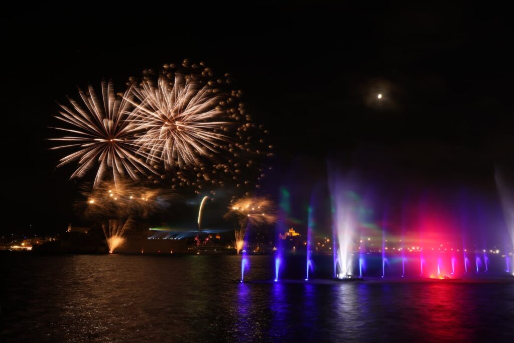 A photo showing Illuminated water fountains and sailing vessels during the Valletta Pageant of the Seas in Malta's Grand Harbour.