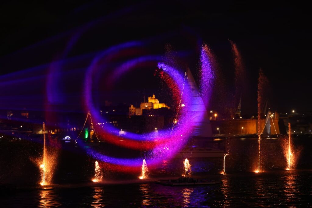 A photo showing Illuminated water fountains and sailing vessels during the Valletta Pageant of the Seas in Malta's Grand Harbour.