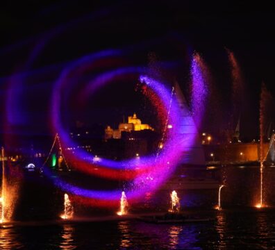 A photo showing Illuminated water fountains and sailing vessels during the Valletta Pageant of the Seas in Malta's Grand Harbour.
