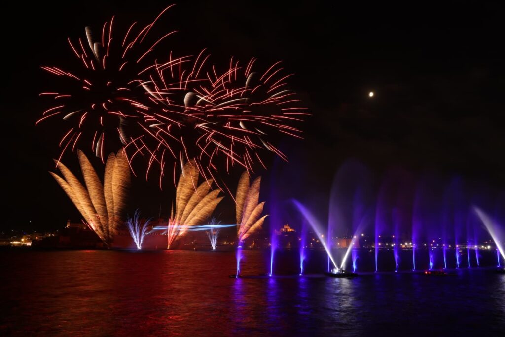 A photo showing Illuminated water fountains and sailing vessels during the Valletta Pageant of the Seas in Malta's Grand Harbour.